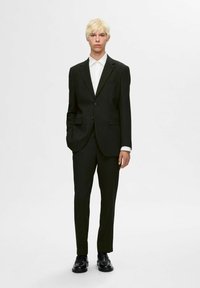 Czarny garnitur dopasowany z gładkiego materiału, z zapięciem na dwa guziki, klapami typu notch i białą koszulą pod spodem. Lśniące czarne buty.