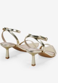 Sandali con tacco color oro dotati di fasce metalliche, schiena aperta e tacco a stiletto basso. Presentano una superficie liscia e lucida e una fibbia regolabile.