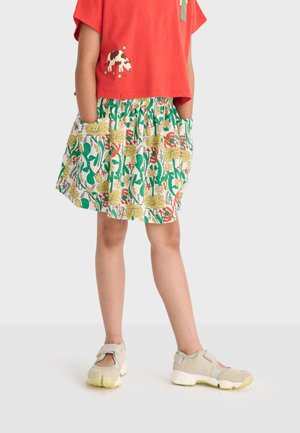 Kind trägt rotes Shirt mit Kuh-Aufnäher, geblümten Rock mit Taschen und beige Sneaker mit gelben Sohlen, steht mit den Händen in den Taschen.