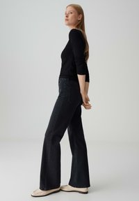 Vrouw met lang blond haar, gekleed in een zwarte lange mouwen top, zwarte flared jeans en witte loafers, staande tegen een effen lichtgrijze achtergrond.
