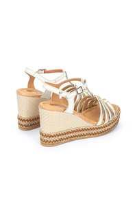 Sandales compensées blanches et dorées avec une texture tressée, présentant plusieurs lanières, une boucle à la cheville et une semelle en jute tressé.