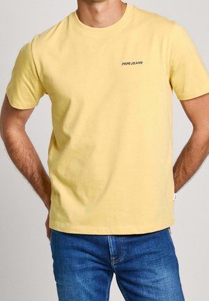 Mann trägt ein gelbes kurzärmeliges Pepe Jeans T-Shirt und blaue Jeans, steht mit den Händen hinter dem Rücken.