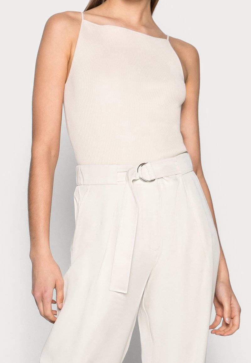 Top con spallina, a coste, di un leggero beige, abbinato a pantaloni bianchi a vita alta con cintura e fibbia ad anello argentato.