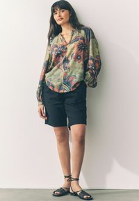 Lichtgewicht blouse met bloemenpatroon in groen, rood en blauw, gecombineerd met zwarte shorts en zwarte sandalen met bandjes op een model.