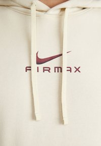 Beige hoodie med en Nike swoosh-logotyp och "AIRMAX"-text i vinröd. Har dragsko-stängningar och slät bomullstyg.