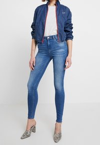 Jeansjacka med röd dragkedja, grå t-shirt, blå skinny jeans och muleklackar med ormskinsmönster. Avslappnad outfit med figurnära silhuett.