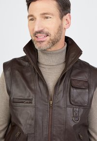 Braune Lederweste mit hohem Kragen, mehreren Taschen und Reißverschluss vorne. Strukturierte Oberfläche mit einem dezenten Glanz und bronze-farbenen Hardware-Details.