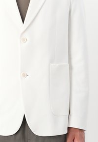 Blazer bianco su misura con texture morbida, design monopetto, due bottoni, revers a punta e tasca laterale.