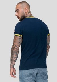 Muž s krátkými vlasy a tetováním na krku a rukou, oblečený v námořnickém tričku se žlutými pruhy na rukávech a světlými modrými džíny, stojí zády.