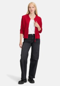 Czerwony sweter z dzianiny, z długimi rękawami, prążkowanymi mankietami i kieszeniami po bokach, nałożony na białą koszulę, zestawiony z czarnymi spodniami o szerokich nogawkach.