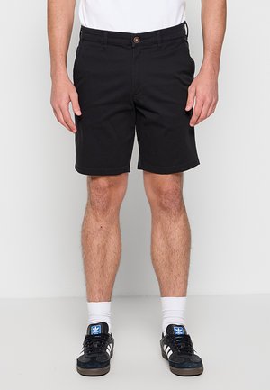 Mężczyzna ubrany w czarne szorty, białe skarpetki, czarne buty sportowe Adidas i białą koszulkę, stojący na jednolitym białym tle.
