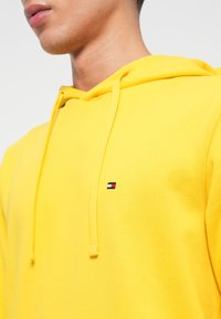 Detailný záber osoby v jasne žltom mikinovom s kapucňou s malou obdĺžnikovou červenou, bielou a modrou nálepkou na hrudi.