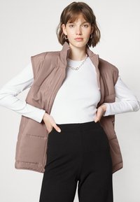 Colete de puffer castanho com gola alta, apresentando um design acolchoado. Usado sobre uma camisa de manga longa branca e combinado com calças pretas.