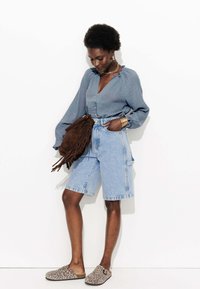Shorts en denim bleu clair avec poches, associés à un haut texturé bleu profond avec un décolleté en V et des détails de boutons. Sac à main à franges marron et sabots à imprimé léopard.