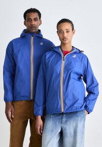 Chaquetas ligeras azules con cremalleras frontales, rayas multicolores y capuchas ajustables, llevadas sobre pantalones casuales.