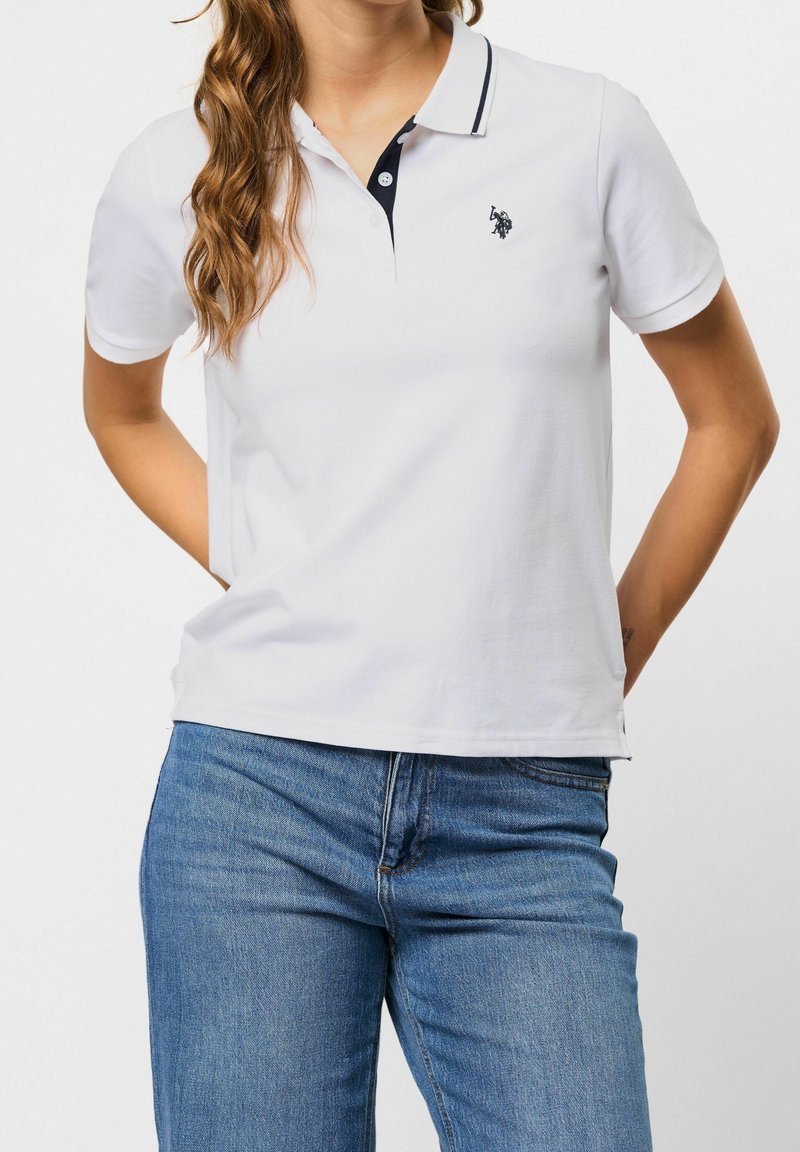 Femme portant un polo blanc à manches courtes avec une bordure noire et un jean bleu, debout les mains dans le dos.