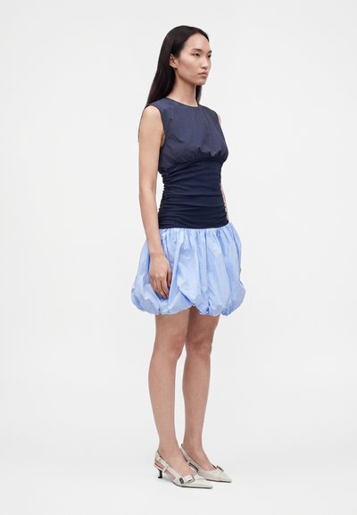 Femme aux longs cheveux foncés portant un haut sans manches bleu marine et une jupe boule bleu clair, debout en talons slingback blancs sur fond uni.
