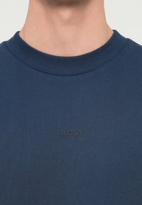 Tmavomodrá rebrovaná mikina s čiernym plastickým logom "BOSS" na prednej strane, okrúhlym výstrihom a textúrovanou látkou.