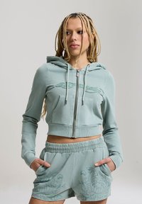 Personne aux longs cheveux tressés porte un sweat à capuche court zippé bleu sarcelle clair et un short assorti à motifs floraux, debout les mains dans les poches.