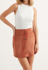 Kostymkjol i orange med fem knappar fram; figursydd siluett och texturerat ribbat mönster. Styld med en vit ribbad linne.