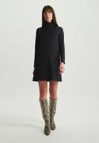 Robe noire sans manches avec un corsage ajusté et une jupe évasée, associée à des bottes en cuir à motif serpent montant jusqu'aux genoux. Texture de tissu lisse.