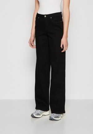 Osoba nosząca czarne jeansy z wysokim stanem i szerokimi nogawkami oraz białe trampki, stojąca z rozluźnionymi ramionami na jasnym, prostym tle.