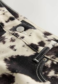Mustvalge laikadega denim püksid, millel on hõbedane nupp, kontrastses õmbluses ja sile tekstuur. Nupule lähedal musta kangast aktsendid.