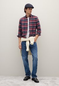 Rotes und marineblaues kariertes Hemd zum Knöpfen über einem weißen T-Shirt, hellblaue Jeans, cremefarbener Pullover um die Taille gebunden und schwarze Lederschuhe; trägt eine marineblaue Mütze.