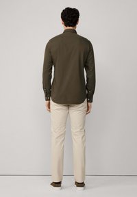 Camisa abotonada verde oliva oscuro de manga larga, combinada con pantalones beige y calzado marrón, vista desde la parte posterior contra un fondo neutro.