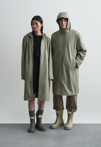 Kaks figuuri kannavad ühesuguseid oliivirohelisi vihmamantleid kapuutsiga. Nad on need koos roheliste kummisaabaste ja tekstureeritud sokkidega, seistes neutraalses taustas.