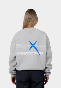 Gai pelēks bērniņu krekls ar pārmērīgu piegriezumu, aizmugurē ir zils grafisks teksts. Mīksta kokvilnas līdzīga tekstūra un ribināti piedurknes.