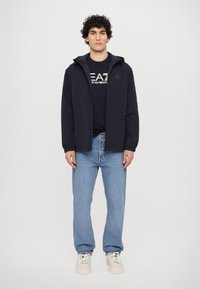 Navy hettejakke, lett stoff, glidelås foran, elastiske mansjetter. Brukt over en svart t-skjorte med hvit grafisk trykk og blå jeans.