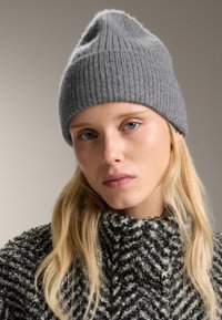 Młoda kobieta z blond włosami, ubrana w szary wełniany beret i czarno-białą sweterkową golf, na beżowym tle.