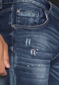 Jeans in denim blu scuro con un design sfilacciato, caratterizzati da diverse aree sfilacciate, macchie di vernice bianche e arancioni e una tasca posteriore.