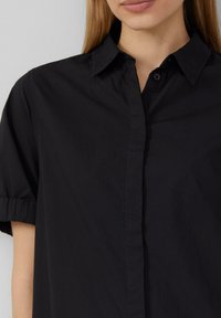 Camicia nera con design a bottoni, maniche corte arrotolate e una consistenza morbida. Presenta un colletto classico e una semplice pattina frontale.