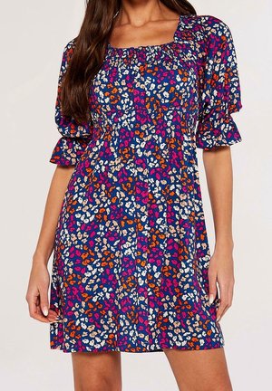 Femme portant une courte robe bleue à manches bouffantes avec un motif floral coloré comprenant des accents orange, rose, blanc et beige.