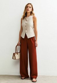 Top senza maniche beige con cinque bottoni marroni, abbinato a pantaloni a gamba larga di color ruggine. Accessoriato con una borsa piccola.