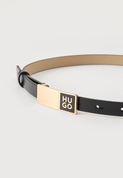 Ceinture en cuir noir avec une boucle rectangulaire dorée portant le mot "HUGO" en lettres noires et dorées.