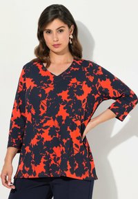 Top floreale a V in rosso e blu navy. Presenta maniche a ¾, una vestibilità ampia e un motivo floreale audace su uno sfondo a contrasto.