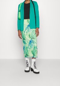 Tyrkysový blejzr přes černý crop top, doplněný zelenou vzorovanou sukní a bílými šněrovacími botami s černou podešví.