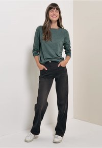 Grünes langärmliges Oberteil, kombiniert mit schwarzen geraden Hosen. Das Modell steht mit den Händen in den Taschen und trägt weiße Sneakers. Glatte Stofftextur.