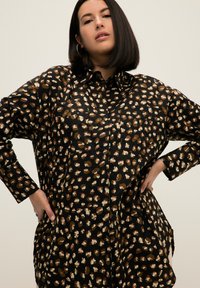 Camicia nera con una fantasia a macchie di leopardo in marrone, crema e verde. Presenta un colletto con bottoni e tasche frontali. Design ampio.