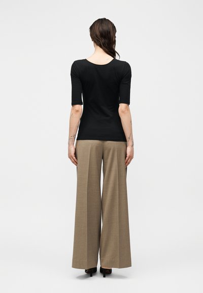 Άτομο με την πλάτη γυρισμένη φοράει εφαρμοστή μαύρη μπλούζα με μανίκια μέχρι τον αγκώνα και μπεζ φαρδιά παντελόνια με μαύρες γόβες.