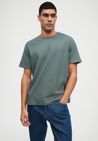 Πράσινο βαμβακερό μπλουζάκι με λαιμόκοψη τύπου crew, κοντά μανίκια και χαλαρή γραμμή, σε συνδυασμό με μπλε τζιν. Λείο υλικό και απλός σχεδιασμός.