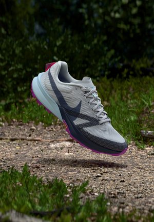 Gaiši pelēka un melna skriešanas apavi ar violetu zoli, kas ir gaisā virs netīrumu takas, fona zaļo lapu ieskauti.