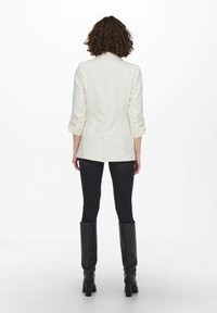 Blazer bianco su misura con maniche arrotolate, spacchetto sul retro, abbinato a jeans skinny neri e stivali neri alti fino al ginocchio, su uno sfondo semplice.
