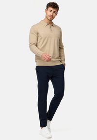 Pull en tricot beige avec un col et trois boutons, associé à un pantalon bleu marine et des baskets blanches pour un look décontracté.