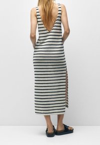 Vrouw in een zwart-wit gestreepte mouwloze maxi-jurk met een diepe V-rug en een zijspleet, gecombineerd met zwarte plateau-sandalen.