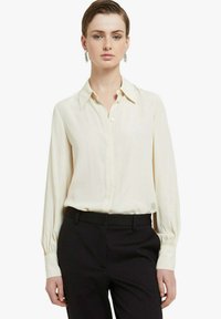 Blusa color crema con colletto, chiusura a bottoni e maniche lunghe. Il tessuto è liscio e leggermente ampio. Abbinata a pantaloni neri.