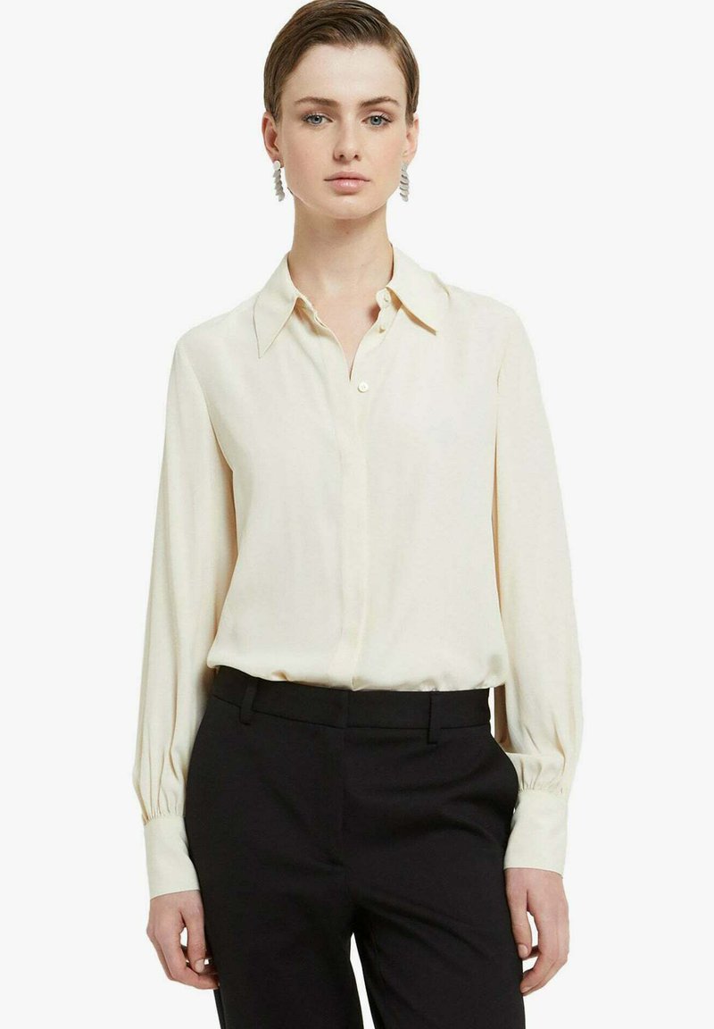 Blusa color crema con colletto, chiusura a bottoni e maniche lunghe. Il tessuto è liscio e leggermente ampio. Abbinata a pantaloni neri.